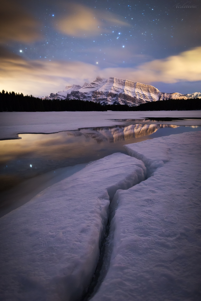 The icy shoreline of Two Jack Lake at night with Mt Rundle and stars reflecting in the small opening in the lake, Banff National Park, Alberta